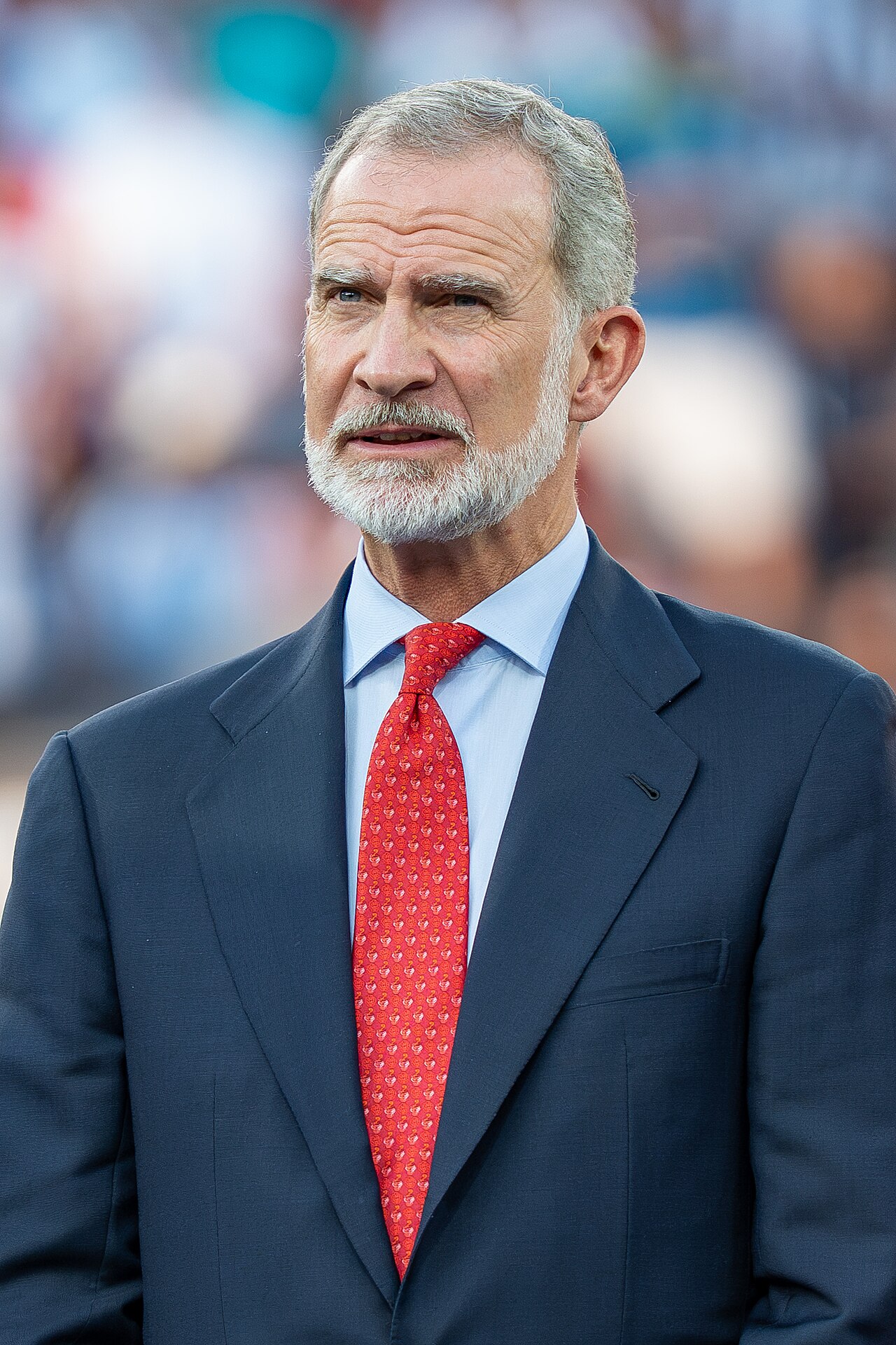 hombre mayor con traje oscuro y corbata roja de patrón, barba gris, mirando hacia un lado; fondo borroso que sugiere multitud o evento al aire libre Fotografía de archivo.
